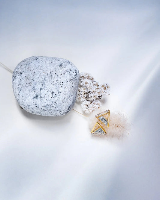 A set of earrings with a geometric diamond-shaped design, featuring silver and gold colors, displayed on a textured grey surface alongside a white flower.